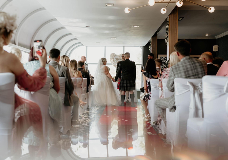 Bride and groom stand at altar holding hands during ceremony, while guests look on from white chairs.